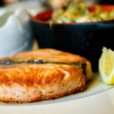A close-up of a cooked salmon steak on a white plate, served with a lemon wedge and a side dish in a black bowl in the background.