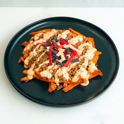 A black plate holds sweet potato fries topped with ground meat, creamy sauce, and garnished with colorful tortilla strips on a white surface.