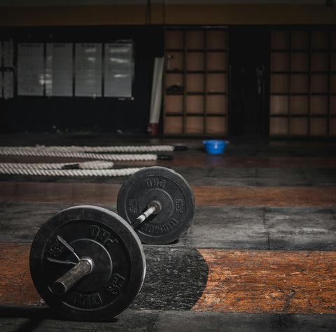 A barbell with black weight plates sits on a worn gym floor, with thick battle ropes coiled in the background and a blue bucket near the wall. The gym appears empty and dimly lit.
