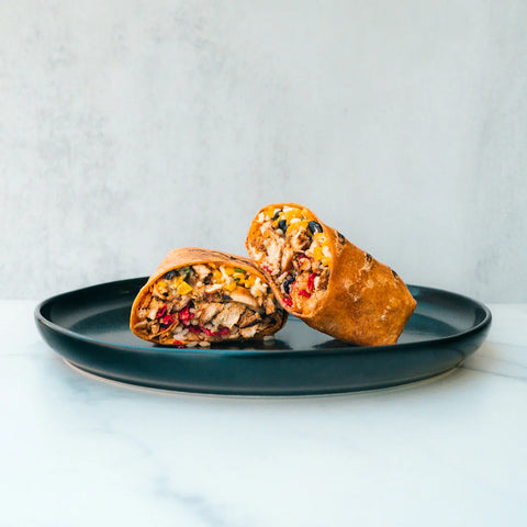 A burrito cut in half, showing a filling of rice, beans, meat, and vegetables, is placed on a dark plate against a light, minimalist background.
