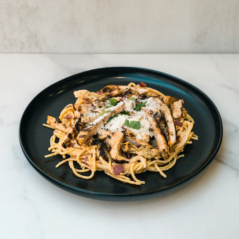 A black plate of spaghetti topped with sliced grilled chicken, garnished with grated cheese and fresh herbs, sits on a white marble surface against a light background.