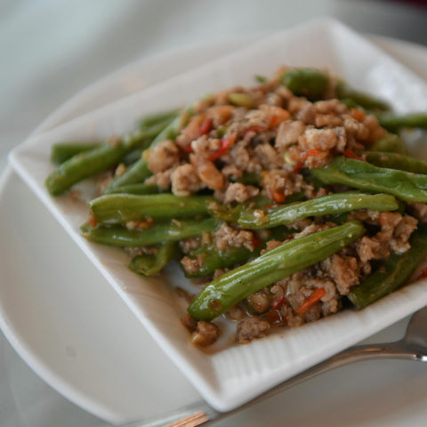 A white plate with stir-fried green beans mixed with ground meat and red chili pieces, served with a spoon on the side.