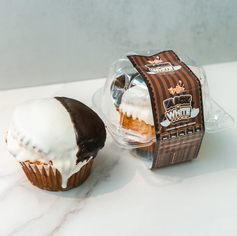 A black and white frosted cupcake sits next to a clear plastic container holding another identical cupcake with a brown striped label that reads Black White Cookie.