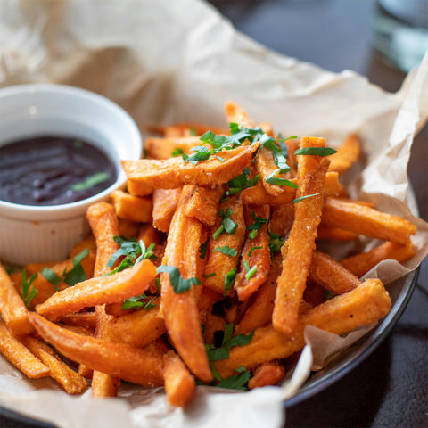 A bowl of sweet potato fries garnished with chopped parsley, served on parchment paper with a small cup of dark dipping sauce on the side.