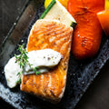 Grilled salmon fillet topped with creamy white sauce and fresh thyme, served with roasted zucchini and red bell pepper on a dark rectangular plate.