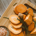 Sliced sweet potatoes on a wooden cutting board with sprigs of fresh rosemary and a small bowl of pink Himalayan salt nearby.