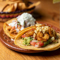 Two shrimp tacos on a decorative plate, topped with grilled shrimp, diced tomatoes, guacamole, corn, and white cheese, with tortilla chips in a bowl in the background.