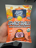 A hand holds a Lesser Evil Space Balls Single Bag, showcasing colorful, space-themed cartoon art and product details on the orange packaging.
