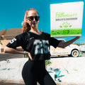 A woman in sunglasses and a Women’s Cropped Mesh Jersey – Black White Trim with the number 25 poses outdoors, holding a large Hummus Fit delivery box on her shoulder, with a vintage truck and building in the background.