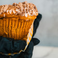 A close-up of a hand in a black glove holding a Basic Baddie Buffin Muffin—pumpkin spice flavor with icing and crumb topping—partially unwrapped from its paper liner, making for a delicious high protein snack.