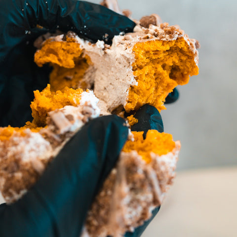 Wearing black gloves, someone pulls apart a crumbly, orange-yellow Basic Baddie Buffin Muffin—a pumpkin spice protein treat dusted with powdered sugar and packed with irresistible flavor for a satisfying, high-protein snack.