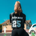 A woman stands outdoors facing away, wearing the Women’s Cropped Mesh Jersey – Black White Trim with "BUFFIN BADDIE 25" on the back. She is in front of a house and a white car under a clear blue sky.