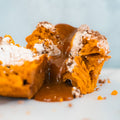 A close-up of a split Basic Baddie Buffin Muffin with orange crumb, dusted with powdered sugar, reveals gooey caramel sauce oozing from the center onto a white surface—an irresistible high protein snack and low caffeine dessert.