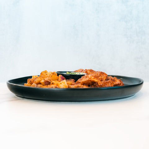 A black plate featuring BBQ Chicken Garlic Parm Potatoes, accompanied by a small container of dipping sauce, is set on a white surface against a light background—a delicious high-protein meal perfect for any occasion.