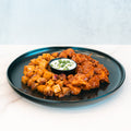 The BBQ Chicken Garlic Parm Potatoes features crispy garlic parmesan potatoes and saucy BBQ chicken pieces for a high-protein meal, served on a black plate with a small cup of creamy herb-topped dipping sauce, all set on a white surface.