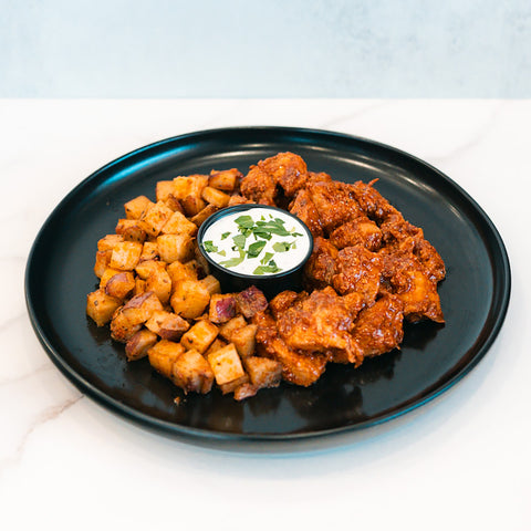 A serving of BBQ Chicken Garlic Parm Potatoes features saucy BBQ chicken pieces for a high-protein meal, roasted Garlic Parmesan Potatoes, and a small cup of white dipping sauce topped with chopped herbs, all presented on a black plate.