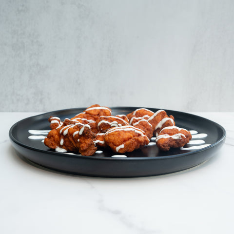 A black plate with pieces of fried chicken, drizzled with white sauce, placed on a white marble surface with a light gray background.