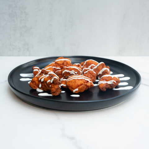 A black plate with pieces of fried chicken drizzled with white sauce, arranged on a white marble surface with a light gray background.