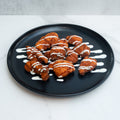 A black plate with pieces of fried chicken drizzled with white ranch sauce, placed on a white marble surface.