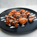A black plate with pieces of fried chicken drizzled with white sauce, arranged on a white surface with extra sauce lines on the plate.