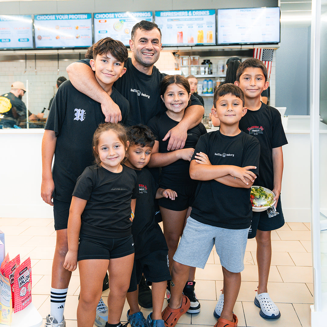 A smiling man stands with six children inside a casual restaurant. They all wear black t-shirts and appear happy, with some of the kids hugging each other. The group poses in front of a counter and menu boards.