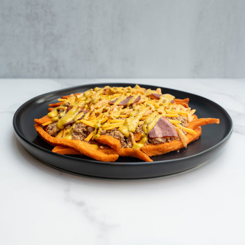 The FitWendy Double Stack Bowl features baked sweet-potato fries topped with ground beef, chopped bacon, cheddar cheese, and yellow sauce—served on a black plate for a high-protein meal against a light gray background.