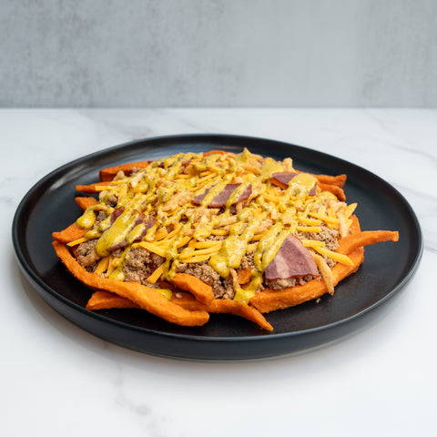 The FitWendy Double Stack Bowl features baked sweet-potato fries with ground beef, shredded cheddar, bacon strips, and yellow sauce for a high-protein meal, served on a black plate over white marble with a gray background.