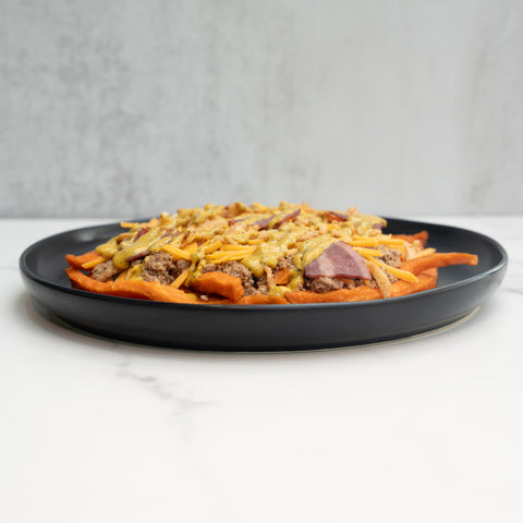 The FitWendy Double Stack Bowl features baked sweet-potato fries topped with high-protein ground beef, melted cheddar cheese, and diced red onions, all served in a black bowl on a white marble surface.