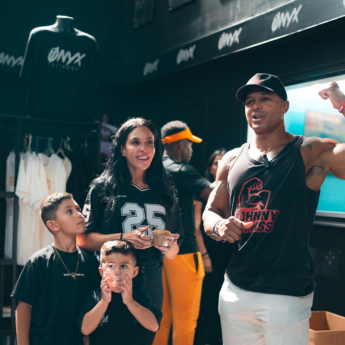 A man in a sleeveless shirt flexes his arm and smiles while standing with a woman and two young boys inside a store. The woman holds a stack of cash, and branded shirts hang in the background.
