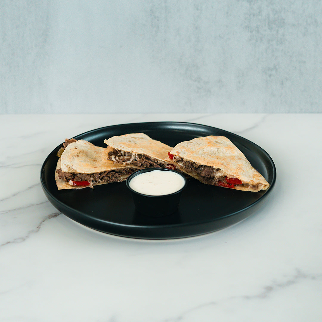 Two wedges of the Philly Cheesesteak Quesadilla, filled with beef and red peppers, are served on a black plate alongside a small cup of creamy white dipping sauce atop a marble surface—making this meal both delicious and macro-friendly.