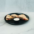 Two wedges of the Philly Cheesesteak Quesadilla, filled with beef and red peppers, are served on a black plate alongside a small cup of creamy white dipping sauce atop a marble surface—making this meal both delicious and macro-friendly.