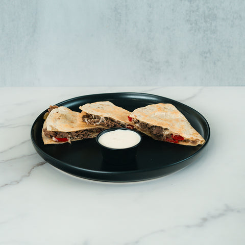 Two wedges of the Philly Cheesesteak Quesadilla, filled with beef and red peppers, are served on a black plate alongside a small cup of creamy white dipping sauce atop a marble surface—making this meal both delicious and macro-friendly.