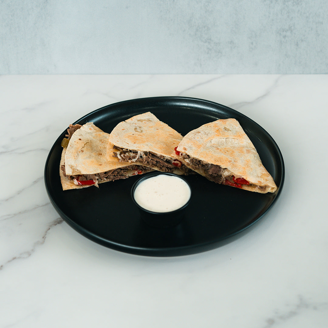 Three Philly Cheesesteak Quesadilla wedges featuring visible beef and red peppers are served on a black plate with a small cup of white dipping sauce, all set atop a white marble surface—perfect for high-protein meal prep.