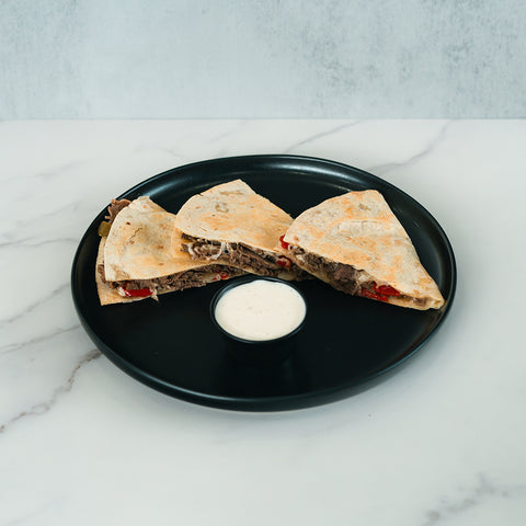 Three Philly Cheesesteak Quesadilla wedges featuring visible beef and red peppers are served on a black plate with a small cup of white dipping sauce, all set atop a white marble surface—perfect for high-protein meal prep.