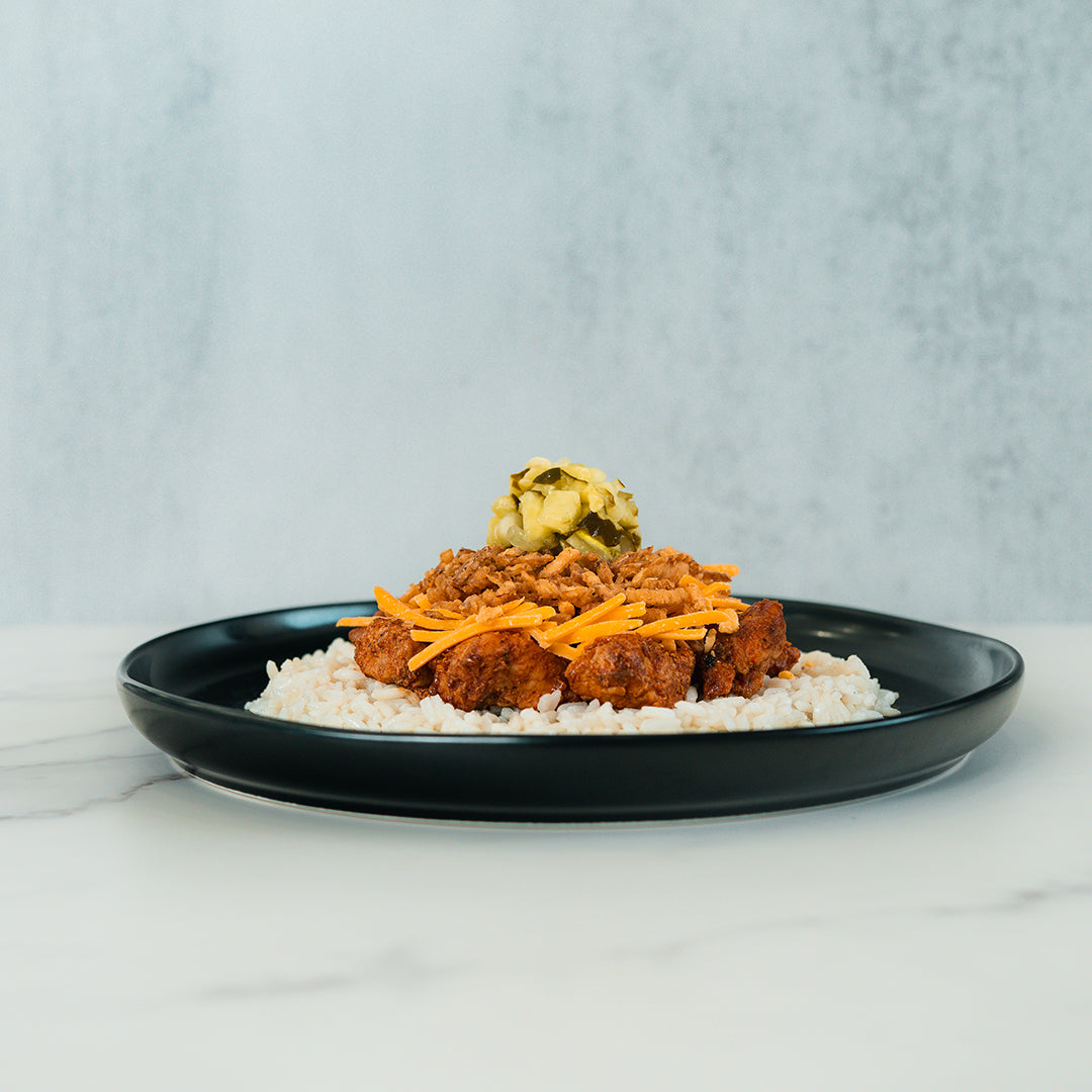 A black plate holds the BBQ Meltdown bowl, featuring rice topped with pieces of fried chicken, shredded cheese, crispy toppings, and a small pile of chopped pickles. Set on a light marble surface against a gray textured background, this high-protein meal is ideal for convenient meal prep.