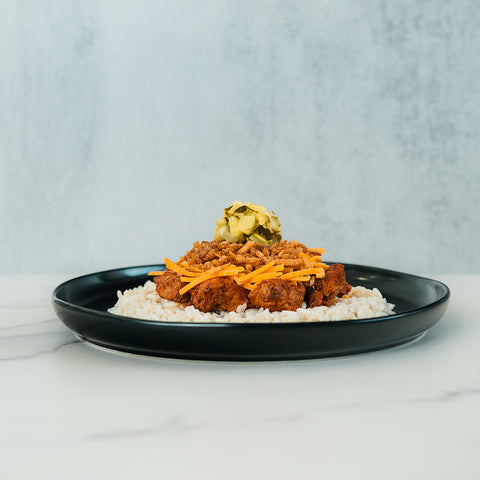 A black plate holds the BBQ Meltdown bowl, featuring rice topped with pieces of fried chicken, shredded cheese, crispy toppings, and a small pile of chopped pickles. Set on a light marble surface against a gray textured background, this high-protein meal is ideal for convenient meal prep.