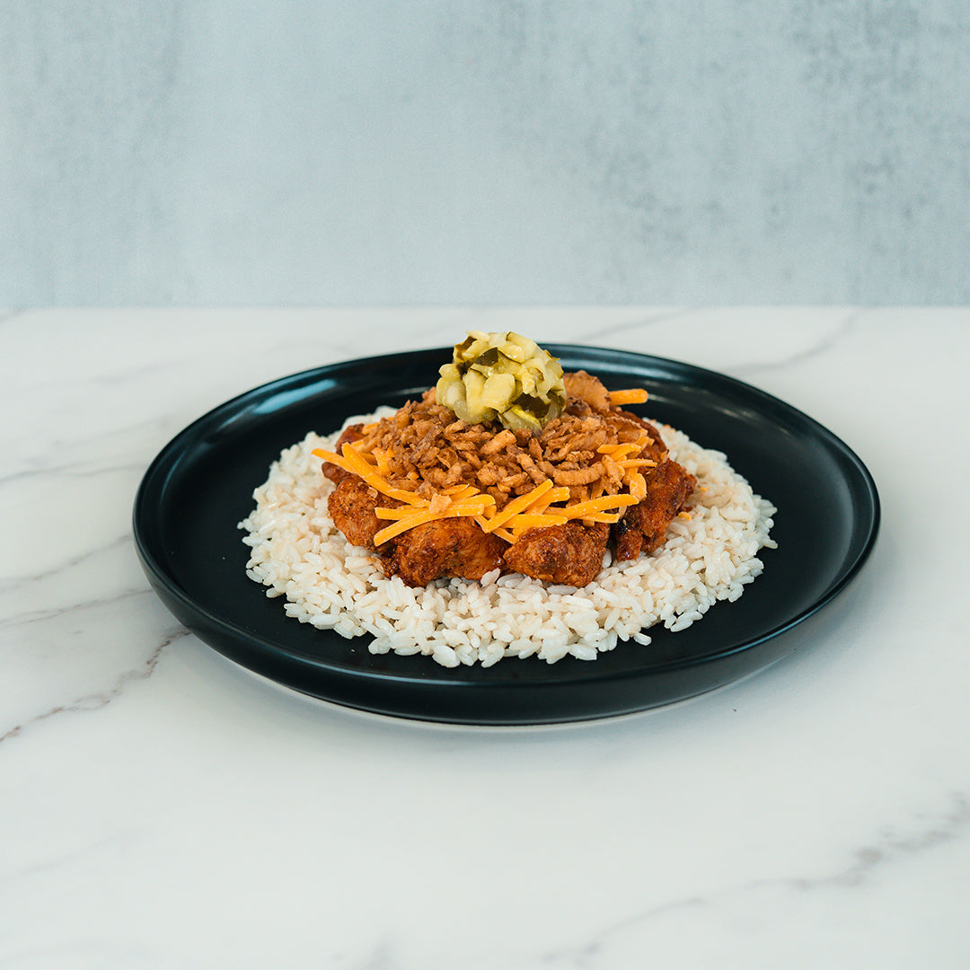 A black plate with white rice topped with BBQ Meltdown, shredded cheddar cheese, and chopped pickles is set on a white marble surface against a light background—a delicious option for healthy meal prep or anyone seeking a high-protein meal prep idea.