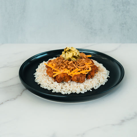 A black plate with white rice topped with BBQ Meltdown, shredded cheddar cheese, and chopped pickles is set on a white marble surface against a light background—a delicious option for healthy meal prep or anyone seeking a high-protein meal prep idea.