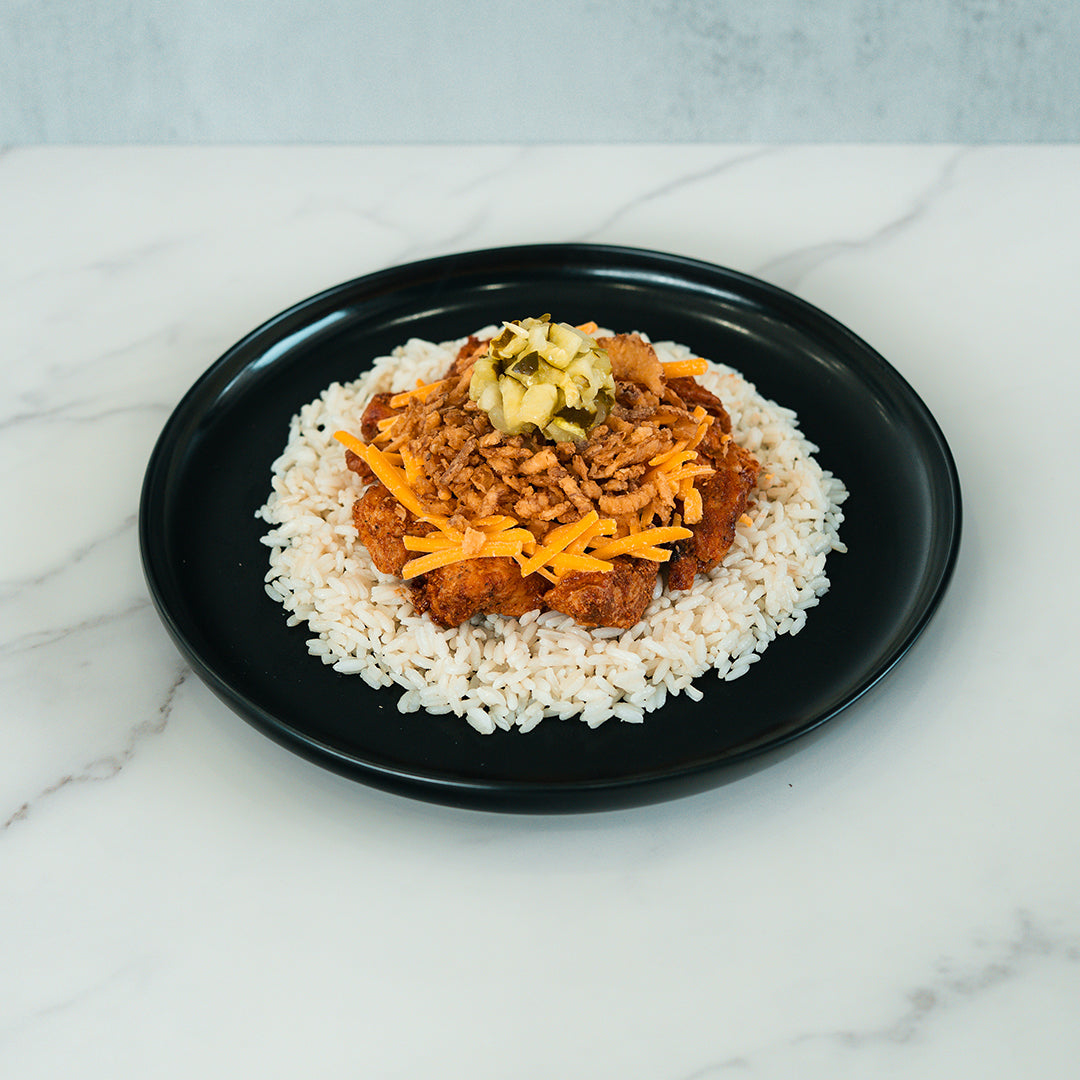 A black plate holds the BBQ Meltdown—a high-protein meal of white rice topped with BBQ chicken, shredded carrots, chopped pickles, and crispy fried onions—creating a colorful dish set on a marble surface.