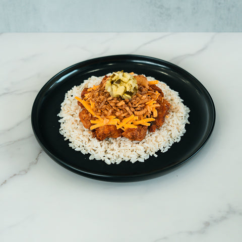 A black plate holds the BBQ Meltdown—a high-protein meal of white rice topped with BBQ chicken, shredded carrots, chopped pickles, and crispy fried onions—creating a colorful dish set on a marble surface.