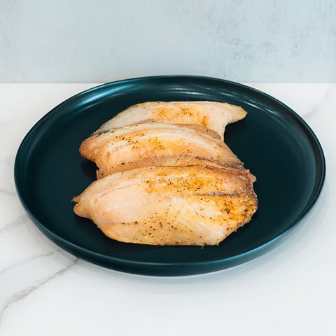A serving of Baked Herbed Tilapia 1lb Competition Approved is arranged on a dark green plate, ideal for meal prep and weight loss, set on a white marble surface against a light gray background.