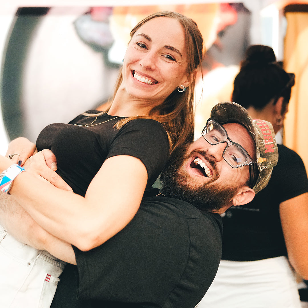 A bearded man in a cap is joyfully holding a smiling woman in his arms. Both appear happy and are wearing casual clothes. Other people are blurred in the background.