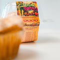 A Basic Baddie Buffin Muffin sits inside a clear plastic container adorned with vibrant, autumn-inspired artwork and nutrition facts. In the foreground, another muffin is softly blurred, drawing attention to this festive and nourishing high-protein snack.