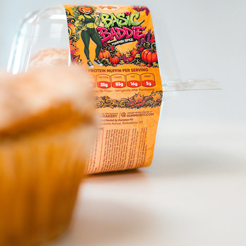 A Basic Baddie Buffin Muffin sits inside a clear plastic container adorned with vibrant, autumn-inspired artwork and nutrition facts. In the foreground, another muffin is softly blurred, drawing attention to this festive and nourishing high-protein snack.