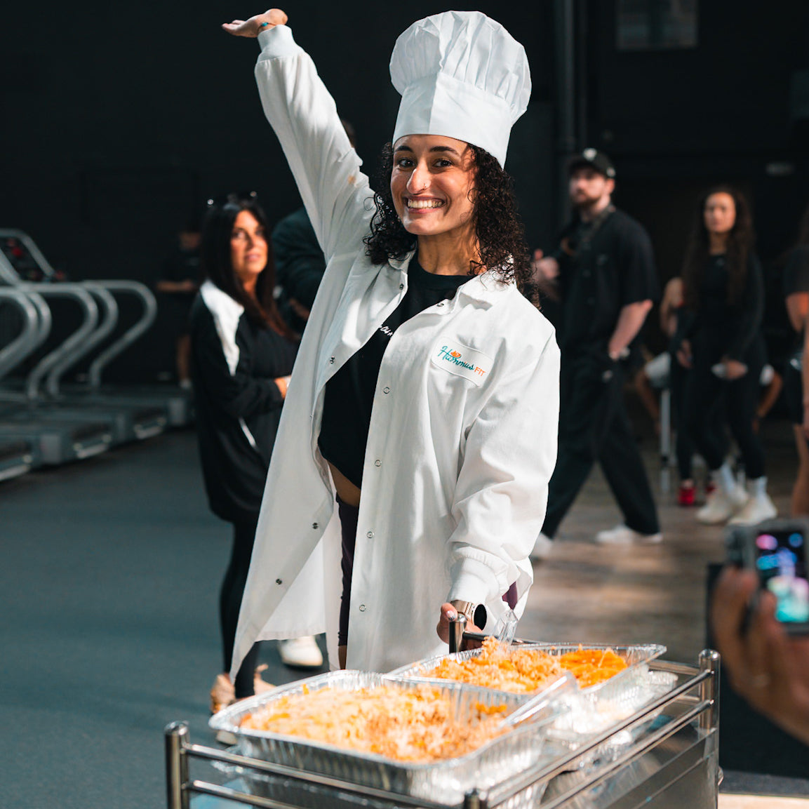 A smiling woman in a chef’s hat and lab coat poses with one arm raised beside a cart holding trays of food. People and exercise equipment are visible in the background.
