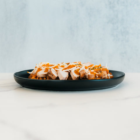 Try this rewritten sentence incorporating the product name: Fit-Fil-A features sliced grilled chicken breast topped with shredded cheese and herbs, served alongside baked waffle fries on a black plate set on a white marble surface against a light blue background—an inviting and healthy dinner option.
