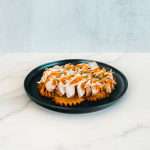 A Fit-Fil-A waffle, topped with sliced smoked chicken breast, shredded cheese, fresh herbs, and a drizzle of sauce, is served on a black plate over white marble—perfect for a healthy dinner.