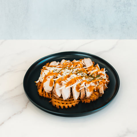 Fit-Fil-A smoked chicken breast, thinly sliced and layered over golden baked waffle fries, is finished with a sprinkle of shredded cheese, savory sauce, and fresh herbs—beautifully presented on a black plate over white marble for a nutritious dinner option.