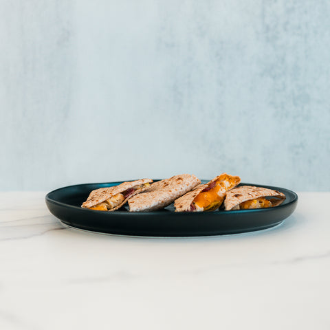 A black plate holds several Strongsville Chicken Ranch Fold quesadillas filled with melted cheese and slices of HALAL Turkey bacon, set on a white marble surface against a light blue background—perfect for anyone seeking a delicious, high protein meal.