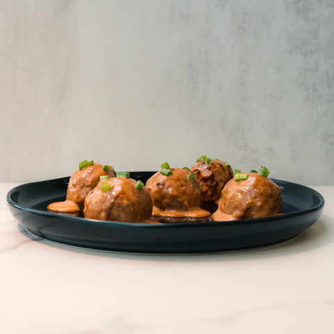 Keto BBQ Chicken Meatballs are served on a black plate, topped with creamy sauce and chopped green onions, creating a high-protein keto meal against a light background.